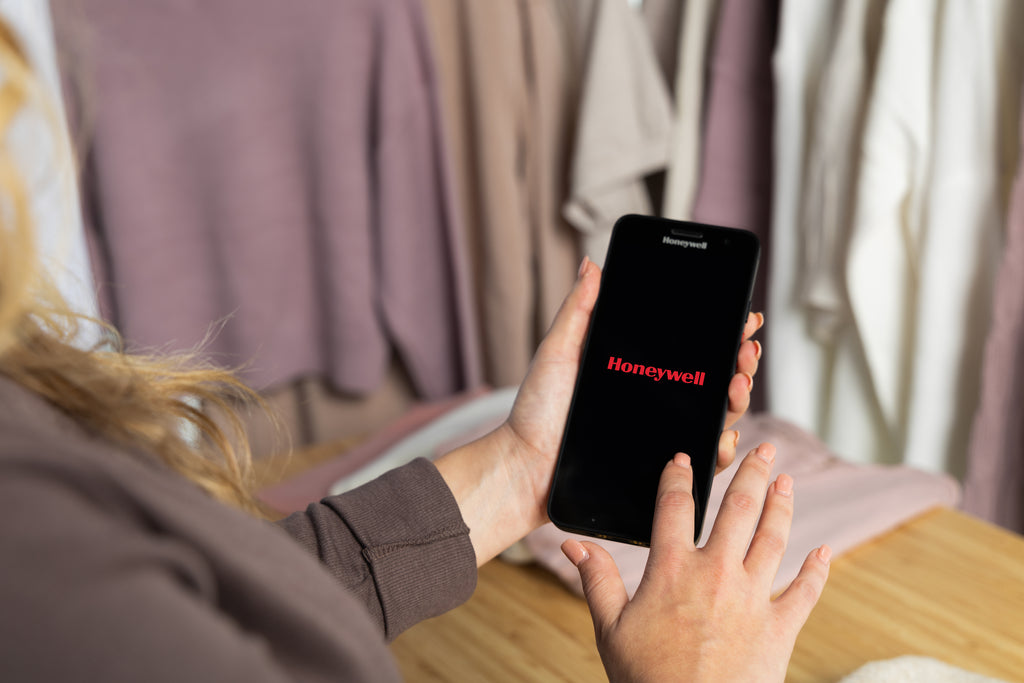 Person holding a smartphone with the Honeywell logo in a room with clothes in the background