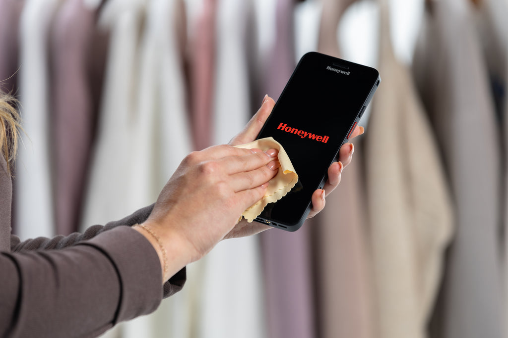 Person cleaning a smartphone with 'Honeywell' logo using a cloth in front of a rack of clothes.
