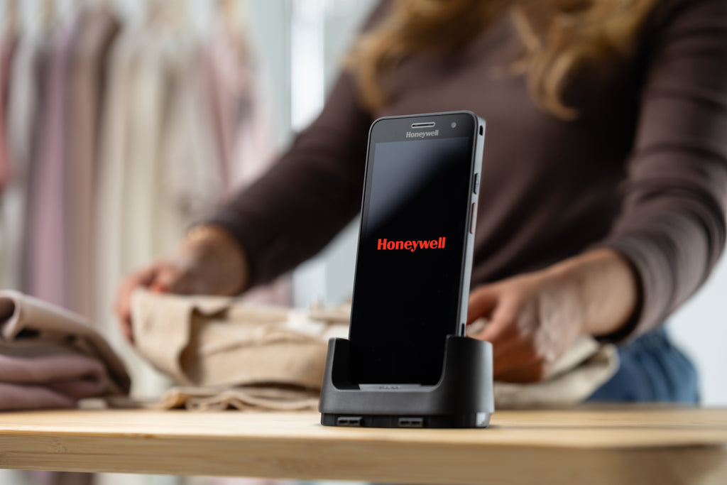 Smartphone on a stand with 'Honeywell' branding on a blurred background of people shopping.