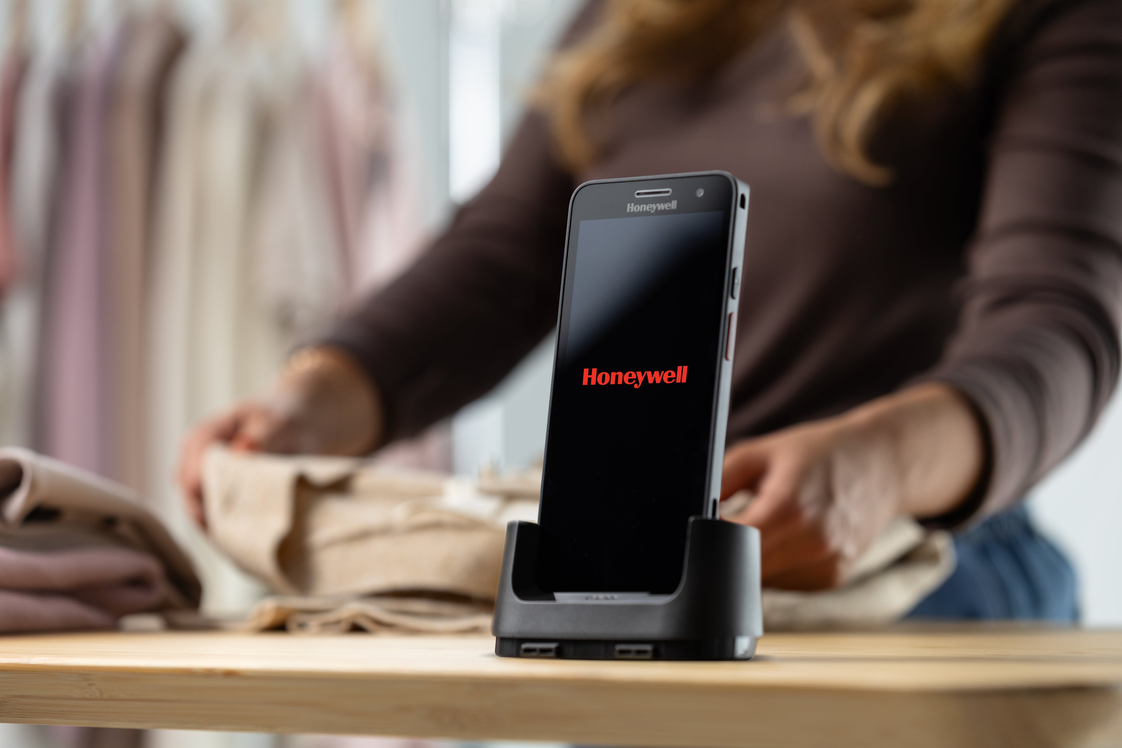 Smartphone on a stand with 'Honeywell' branding on a blurred background of people shopping.