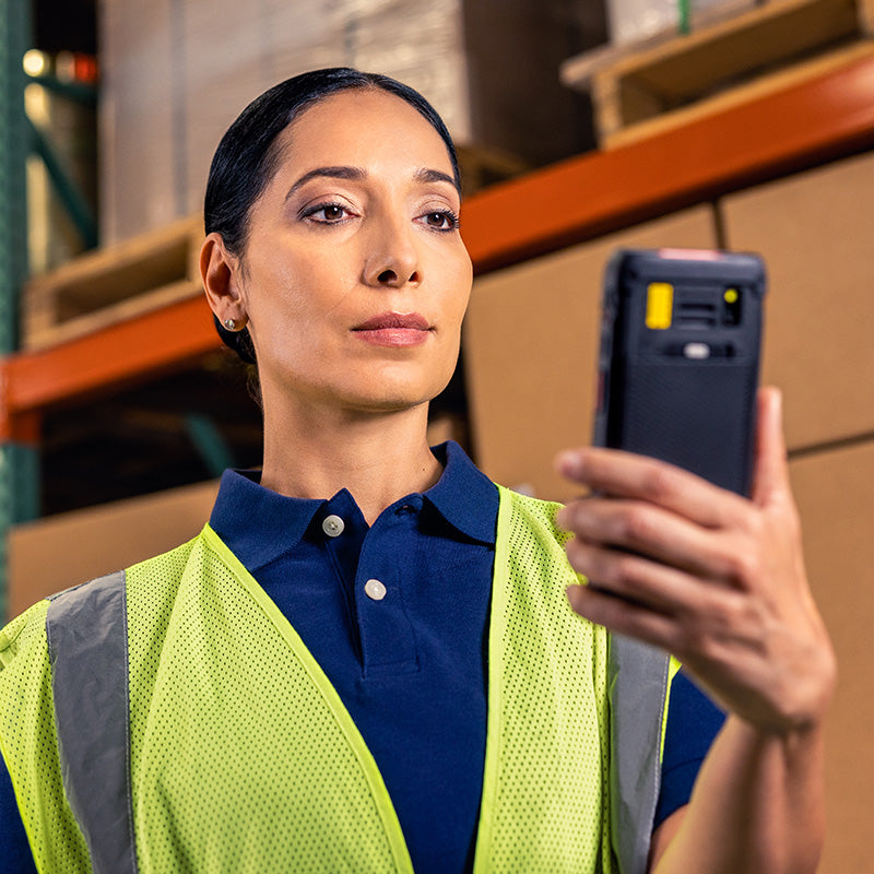 Woman in a warehouse wearing a yellow safety vest and using a handheld device.